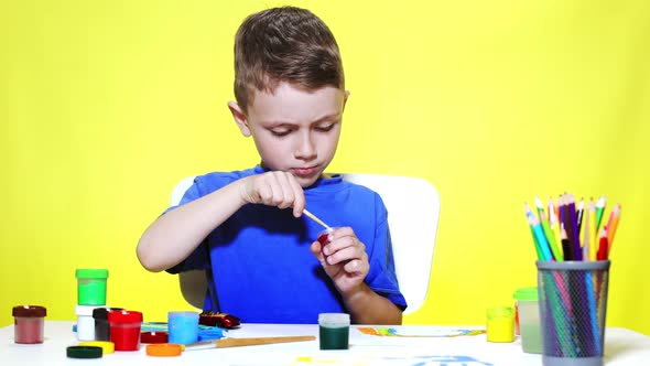 A little preschooler boy is sitting at a table, drawing with watercolor paints on a sheet of paper.