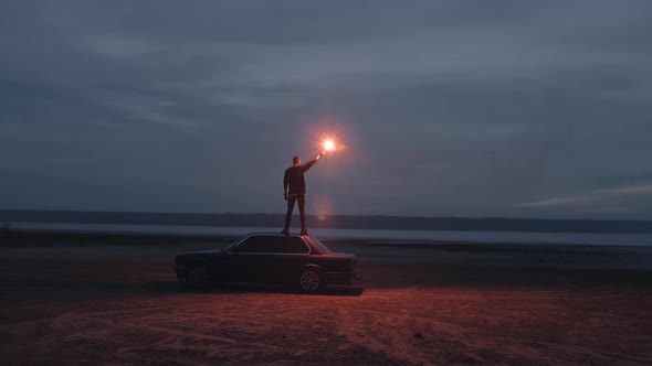 Handheld Cinematic Dark Shot of Young Man Standing on the Car with Red Signal Burning Flare on the