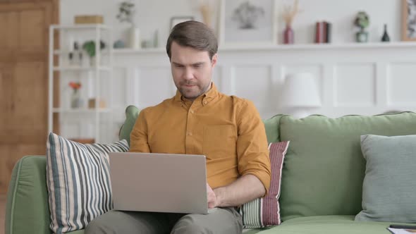 Young Man with Laptop Reacting to Loss on Sofa