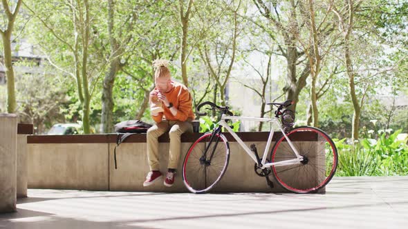 Albino african american man with dreadlocks sitting in park with bike eating sandwich