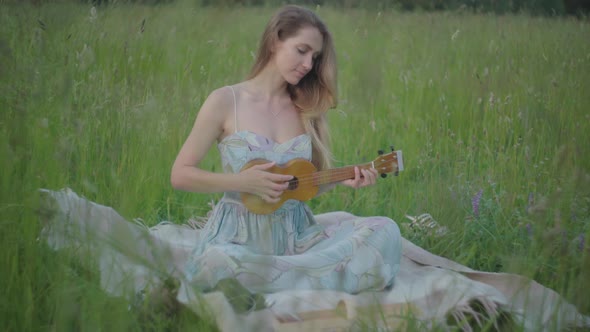 Charming Female Musician Playing Ukulele on Green Meadow. Portrait of Positive Young Caucasian Woman