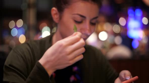 Beautiful Woman Eating Peas While Watching Her Phone at a Restaurant, Smiling Wide