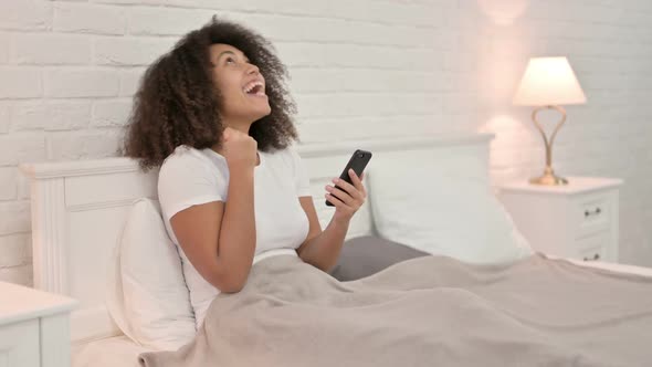 Success Young African Woman Celebrating on Smartphone in Bed