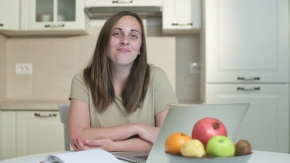 Smiling young woman freelancer with  laptop looks at camera in the kitchen from home