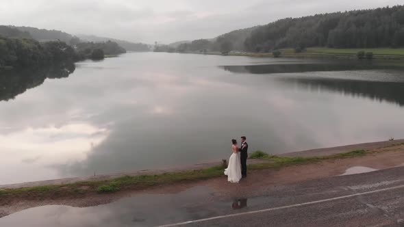 The Bride and Groom Stand Face to Face on the Dam Against the Background of the River and Fog Over