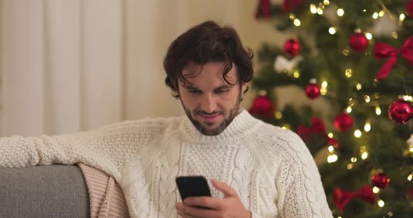 Man Celebration New Year Near Christmas Tree at Home Using Smartphone