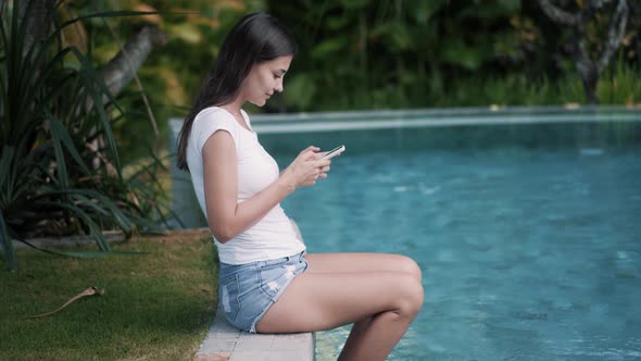 Positive Girl Types on Cellphone Sitting Near Swimming Pool