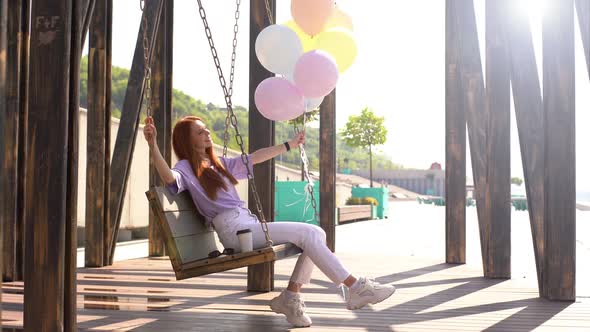 Attractive Redhead Girl Having Fun Carefree Sitting and Swaying on Swing with Fountain of Balloons.