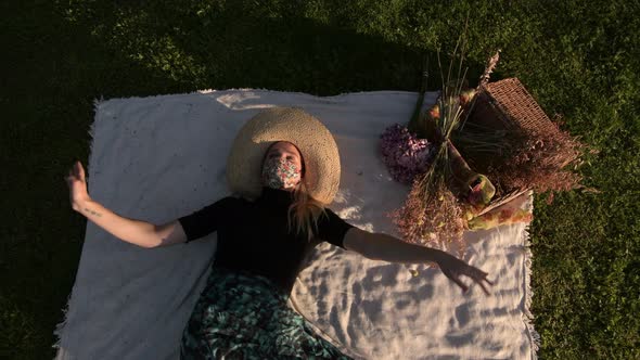 Woman wearing a face mask lying on picnic blanket on the grass waves arms