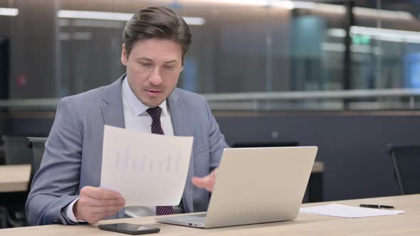 Middle Aged Businessman Reading Documents while using Laptop 