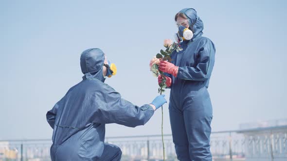 Man Standing on One Knee and Giving Flowers To Woman in Antiviral Suit. Happy Young Caucasian Girl