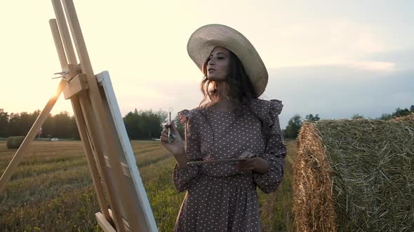 Artist girl paints with brushes on canvas in a field at sunset