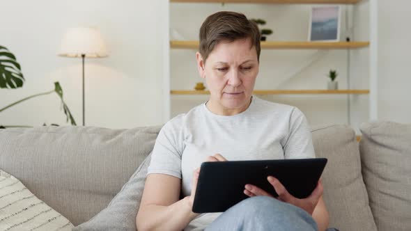 Smiling Elderly Mature Woman Resting on Sofa Using Digital Tablet Alone at Home