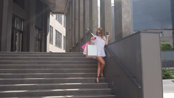 Girl with Phone Staying on Shopping Mall Stairs with Colorful Shopping Bags and Drinking Coffee