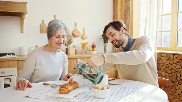 Caring Adult Son Pouring Fresh Hot Tea for His Greyhaired Mother and Him Enjoying Cozy Breakfast and