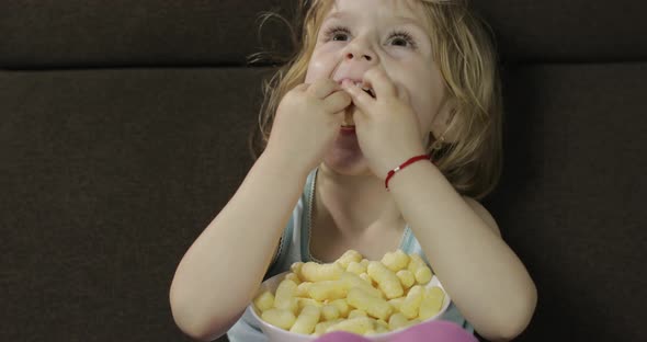 Girl Sitting on Sofa and Eating Corn Puffs. Child Smiling and Taste Puffcorns