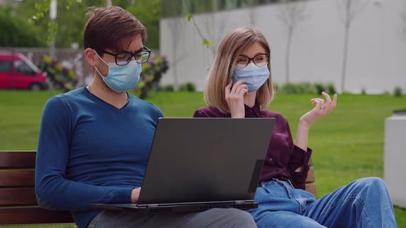 Two professional coworkers in protective mask working sitting on bench spring background.