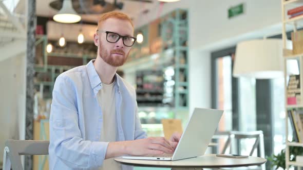 Redhead Man with Laptop Smiling at Camera in Cafe
