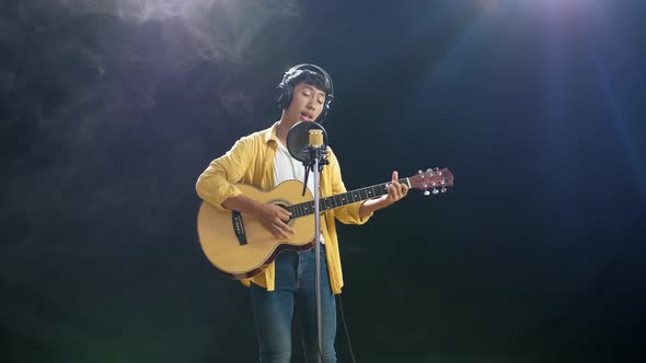 Young Asian Boy With Headphone Playing A Guitar And Singing On The White Smoke Black Background