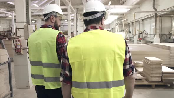 Two Workers Talking in Green Waistcoats and Helmets at a Furniture Factory Among Machines