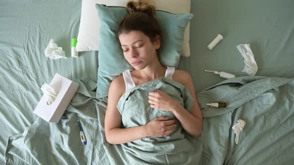 A sick-looking young woman lies and suffers in bed. Medicines are scattered around
