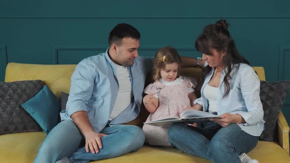 Young Family Reads a Book to Their Little Daughter While Sitting on the Couch