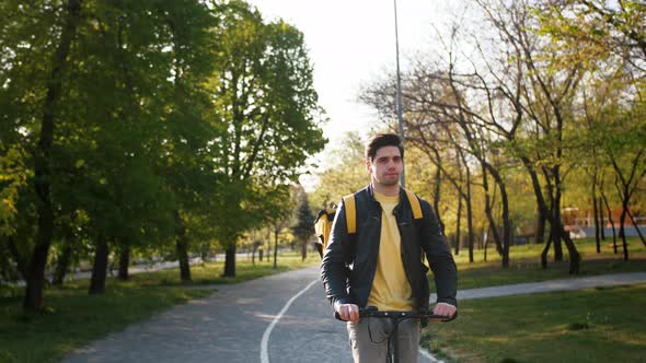 Portrait of Delivery Man with Yellow Bagpack Riding Electric Scooter Through the City Park with Food