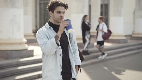 Tattooed Curlyhaired Caucasian Man Drinking Coffee and Smiling at Camera with Blurred Hippie Friends