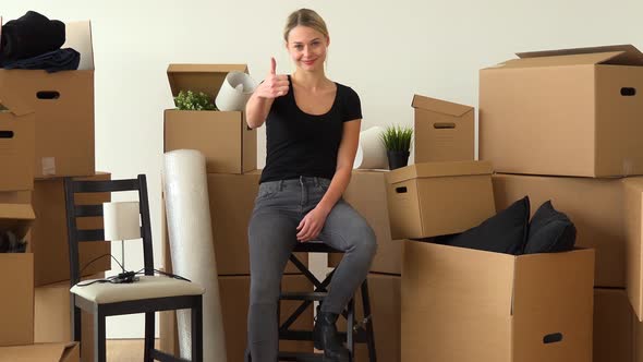 A Happy Moving Woman Sits on a Chair in an Empty Apartment and Shows a Thumb Up To the Camera