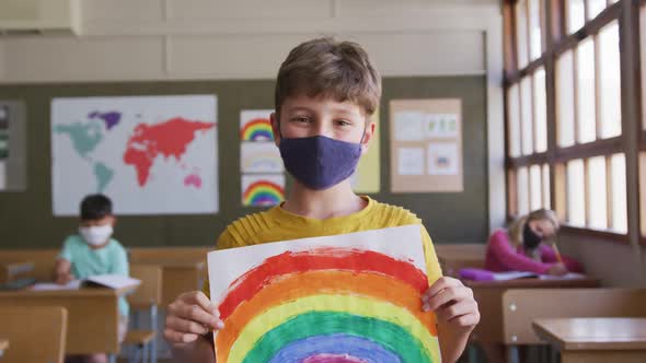 Boy wearing face mask holding a rainbow painting in class at school 