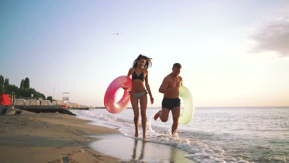 Happy Young Couple Runnung on the Beach with Rubber Rings During Sunset Slow Motion