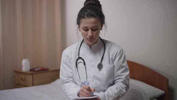 Portrait of Concentrated Young Doctor Sitting on Bed Listening Looking at Camera Writing with Pen