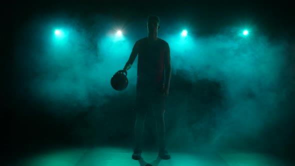Silhouette of a Young Guy Dribbling Basketball on a Dark Studio Background with Smoke and Blue