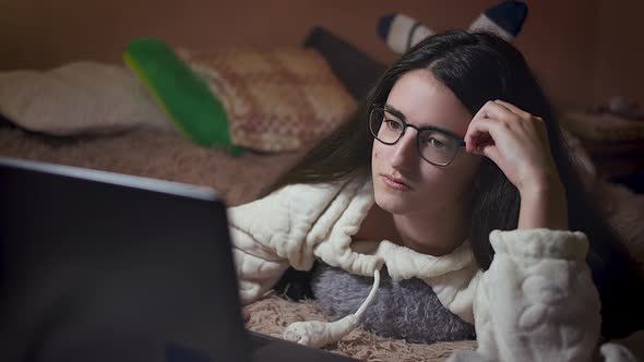 Girl student with glasses prepares homework on a laptop while lying in her bed in the evening