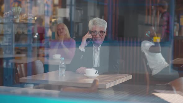 View Through Window of Senior Businessman Have Phone Call Sitting in Coffee Shop