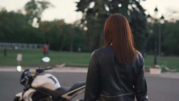 Stylish Young Woman Motorcyclist in a Leather Jacket Goes to Her Bike and Puts on a Helmet