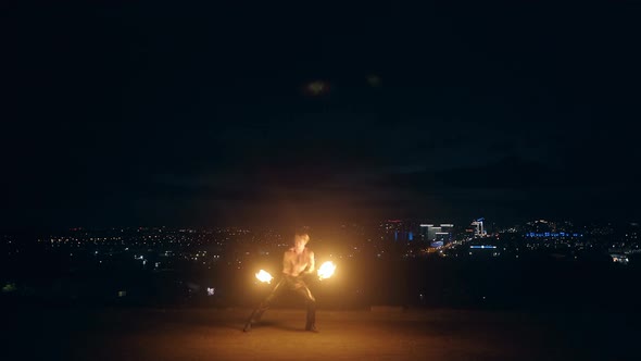 Young Blond Male Does Fire Dance with Two Torches Spins Torches in the Middle of the Night with City