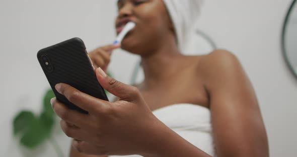 Happy african american woman brushing teeth and using smartphone in bathroom