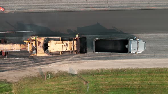 Birds eye view of asphalt paver laying blacktop and dump truck refilling with loose aggregate.