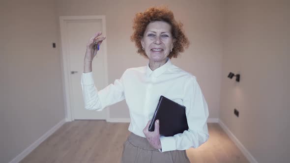 Happy Elderly Woman Realtor Shows the Keys to the Real Estate to the Camera on the Background of an