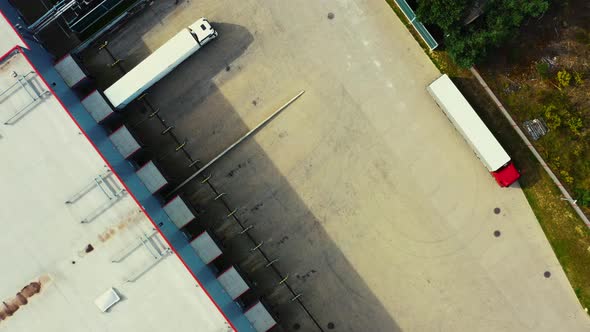 Aerial following shot of semi-trailer trucks travelling along a warehouse on the parking lot in the