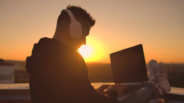A Man Freelancer in Headphones Standing on the Roof at Sunset Writes on the Keyboard Code Pages