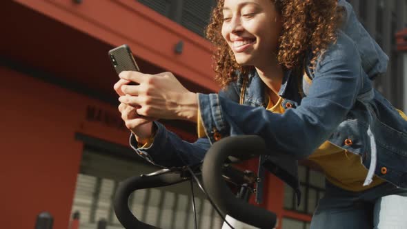 Happy biracial woman in city, sitting on bike using smartphone