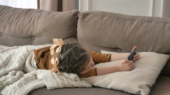 Little Girl Is Lying on the Couch at Home with Her Phone in Her Hands and Watching Cartoons