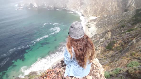 A young woman sitting on top of a cliff with the Bixby Creek Bridge in the background.