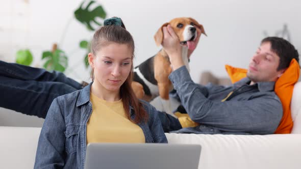 Woman Working with Laptop While Man Lying on Couch with Dog in Home Room Spbi