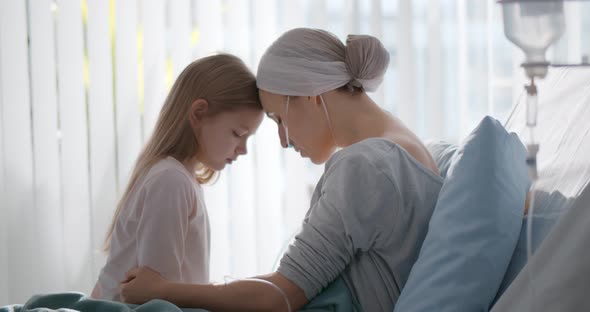Side View of Young Woman with Cancer Sitting on Hospital Bed with Little Daughter