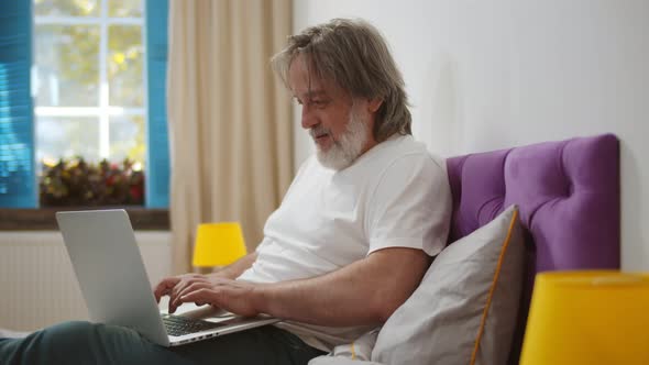 Senior Man Sitting on Bed Working Online From Home with Laptop