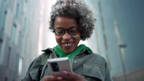 Smiling adult African woman wearing casual clothes texting by phone near buildings