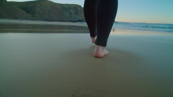 Camera Follows Barefoot Surfer in Wetsuit on Beach